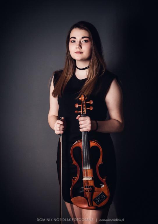 Portret młodej kobiety w czarnej sukience, trzymającej skrzypce elektryczne i smyczek na ciemnoszarym tle.