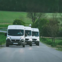 Kolumna trzech białych busów (Fiat Ducato i Mercedes Sprinter) jadących po asfaltowej drodze przez zielony krajobraz, widok z przodu.