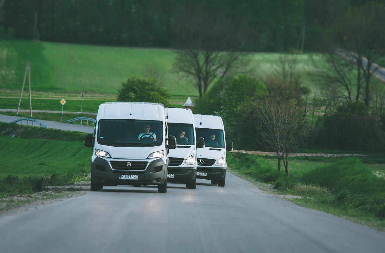 Kolumna trzech białych busów (Fiat Ducato i Mercedes Sprinter) jadących po asfaltowej drodze przez zielony krajobraz, widok z przodu.