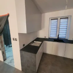 Nowoczesna kuchnia w szaro-białej tonacji z czarnym blatem i zlewozmywakiem, widok z perspektywy wejścia. W tle okno z żaluzjami i fragment łazienki w trakcie remontu.