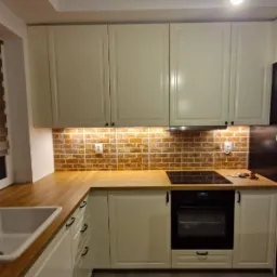 Jasna kuchnia na wymiar w Radomiu: kremowe szafki z drewnianym blatem, ceglana ściana nad blatem, czarna płyta indukcyjna i piekarnik. Oświetlenie podszafkowe.