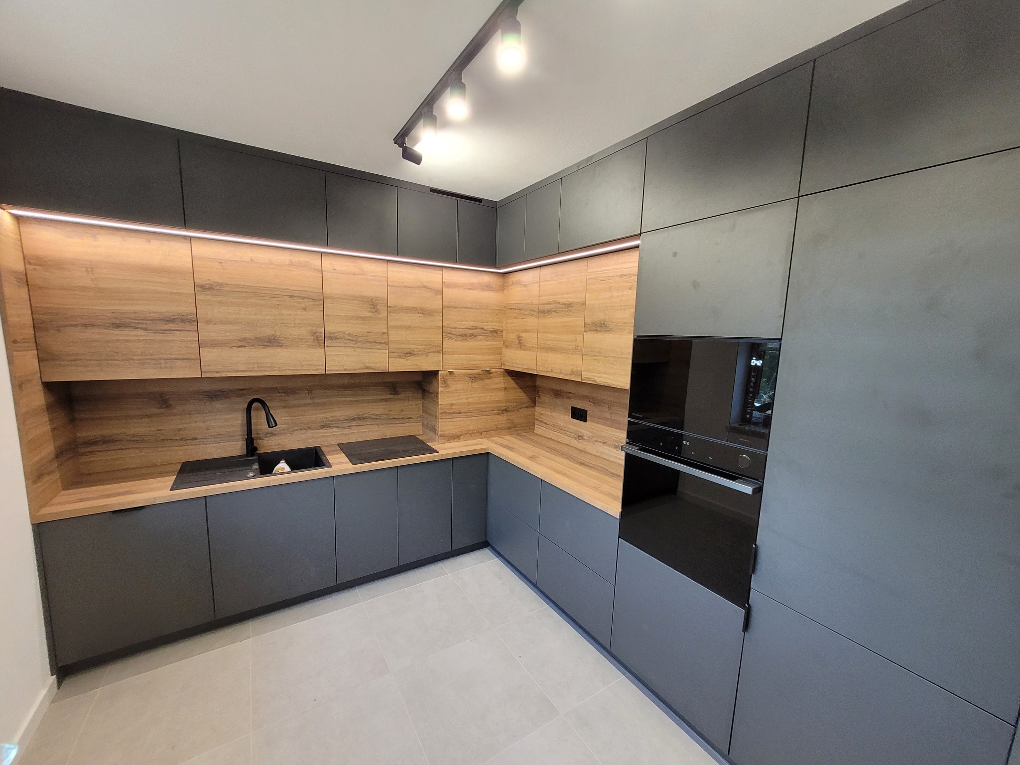 Nowoczesna kuchnia na wymiar z szarymi szafkami dolnymi i drewnopodobnymi górnymi, oświetlona listwą LED i reflektorami, z czarnym zlewem i płytą.