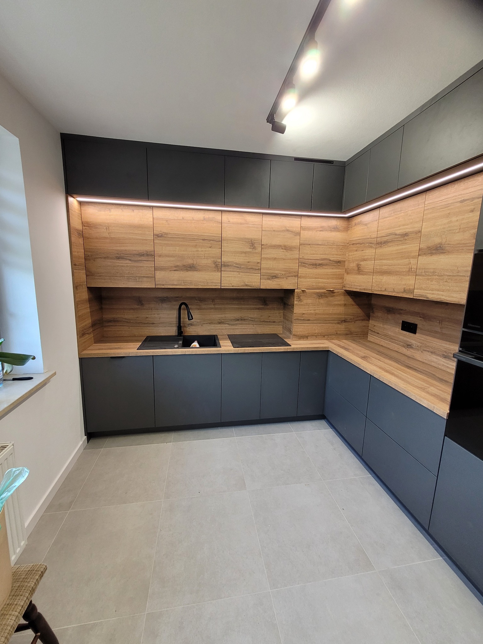 Nowoczesna kuchnia z drewnianymi frontami szafek górnych i grafitowymi dolnymi, oświetlona ledami. Widoczny czarny zlew i płyta grzewcza.