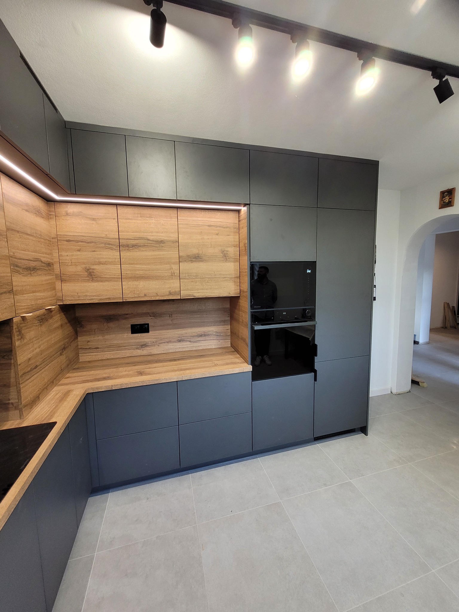 Nowoczesna kuchnia: szare szafki dolne, drewnopodobne górne z oświetleniem LED. Na ścianie wbudowany piekarnik. Podłoga wyłożona jasnymi płytkami.