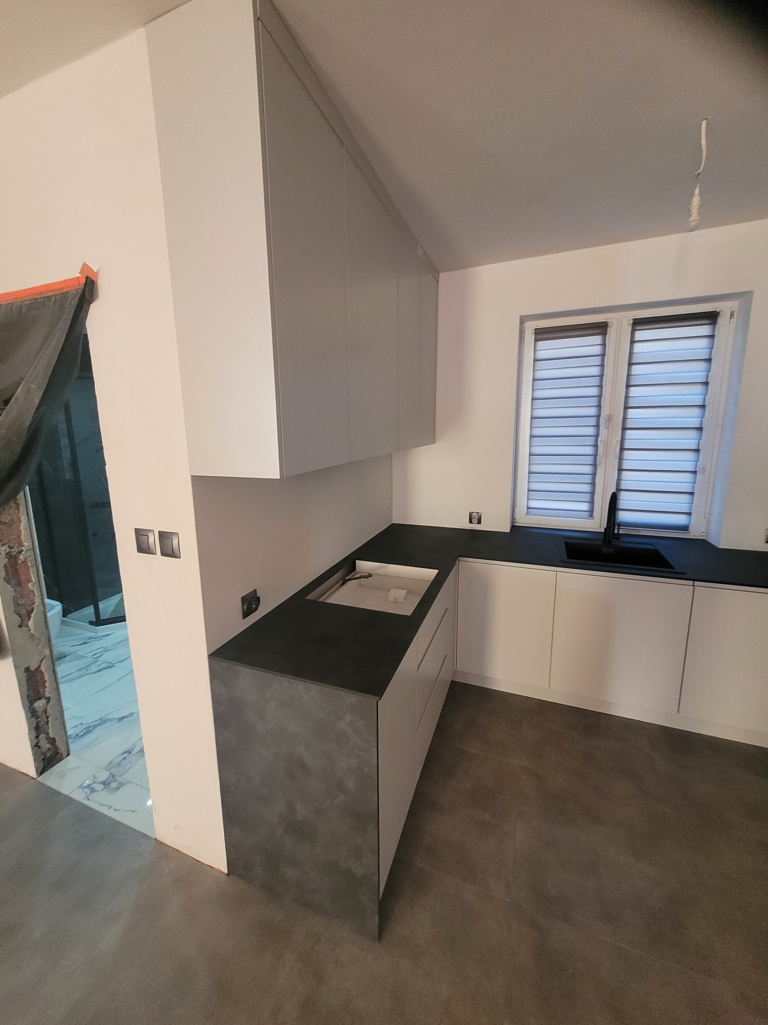 Nowoczesna kuchnia w szaro-białej tonacji z czarnym blatem i zlewozmywakiem, widok z perspektywy wejścia. W tle okno z żaluzjami i fragment łazienki w trakcie remontu.