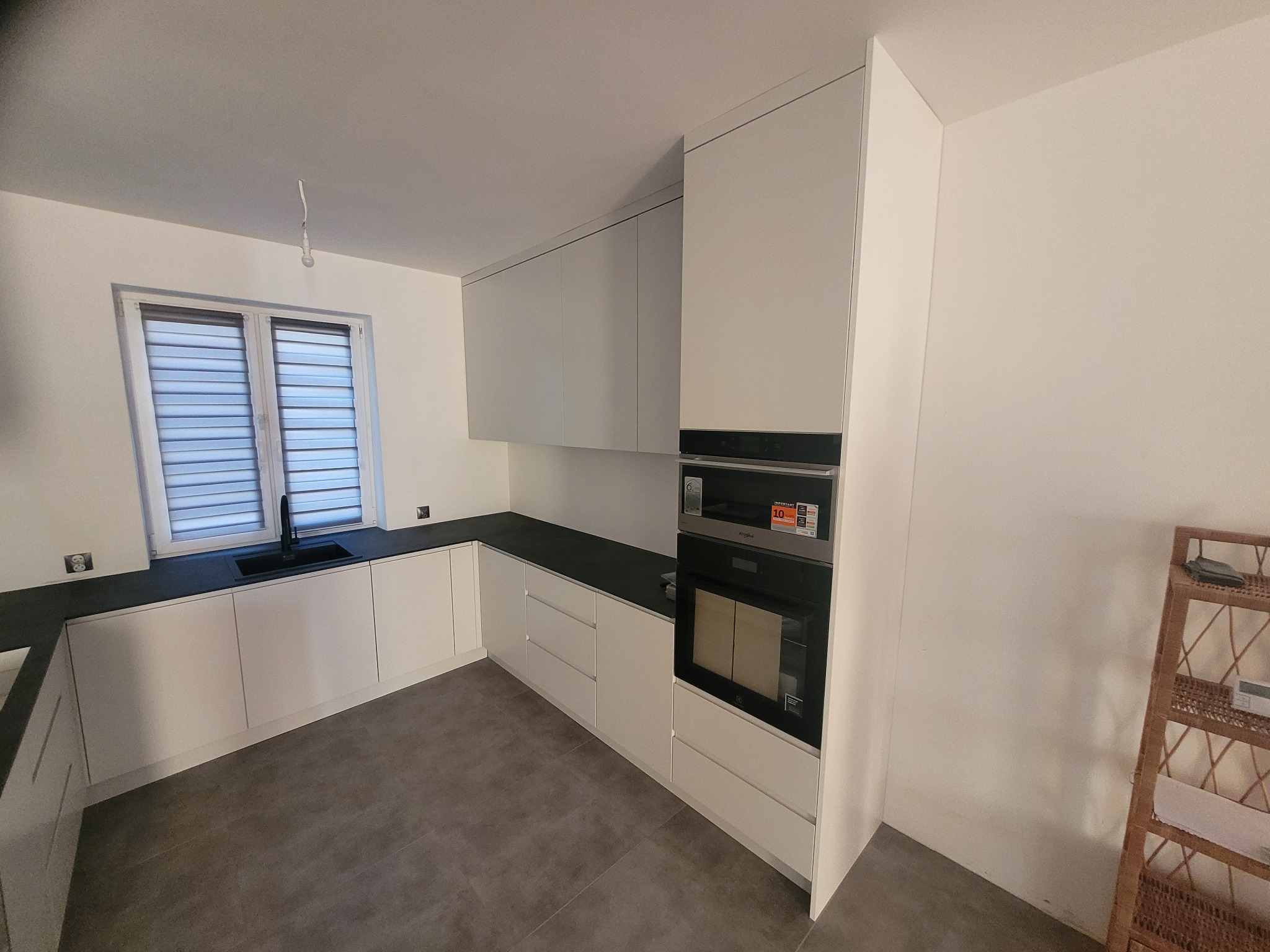 Nowoczesna, minimalistyczna kuchnia z białymi frontami i czarnym blatem, oświetlona światłem dziennym wpadającym przez okno z roletami.