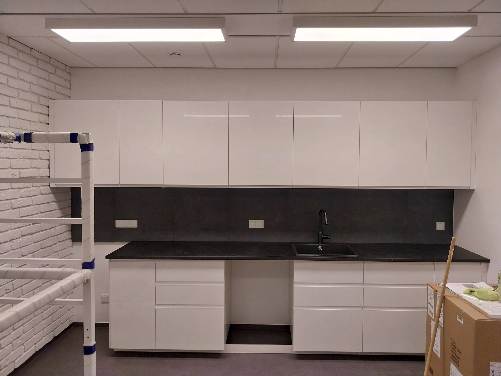 Biała, nowoczesna zabudowa kuchenna z czarnym blatem i panelem ściennym. Minimalistyczny design, oświetlenie LED, fragment rusztowania malarskiego po lewej.