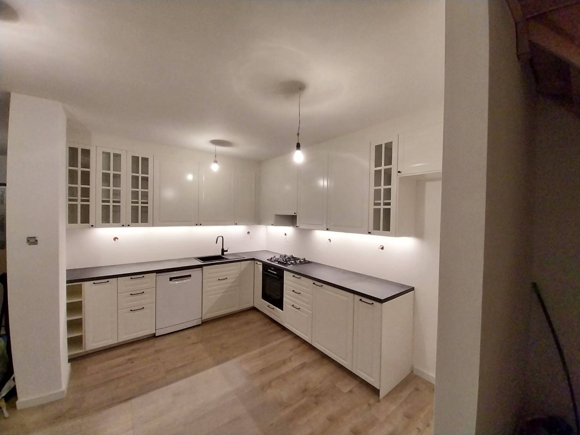 Jasna kuchnia z białymi szafkami i ciemnym blatem, oświetlona wiszącymi lampami i światłem spod szafek. Widoczna płyta gazowa i drewniana podłoga.