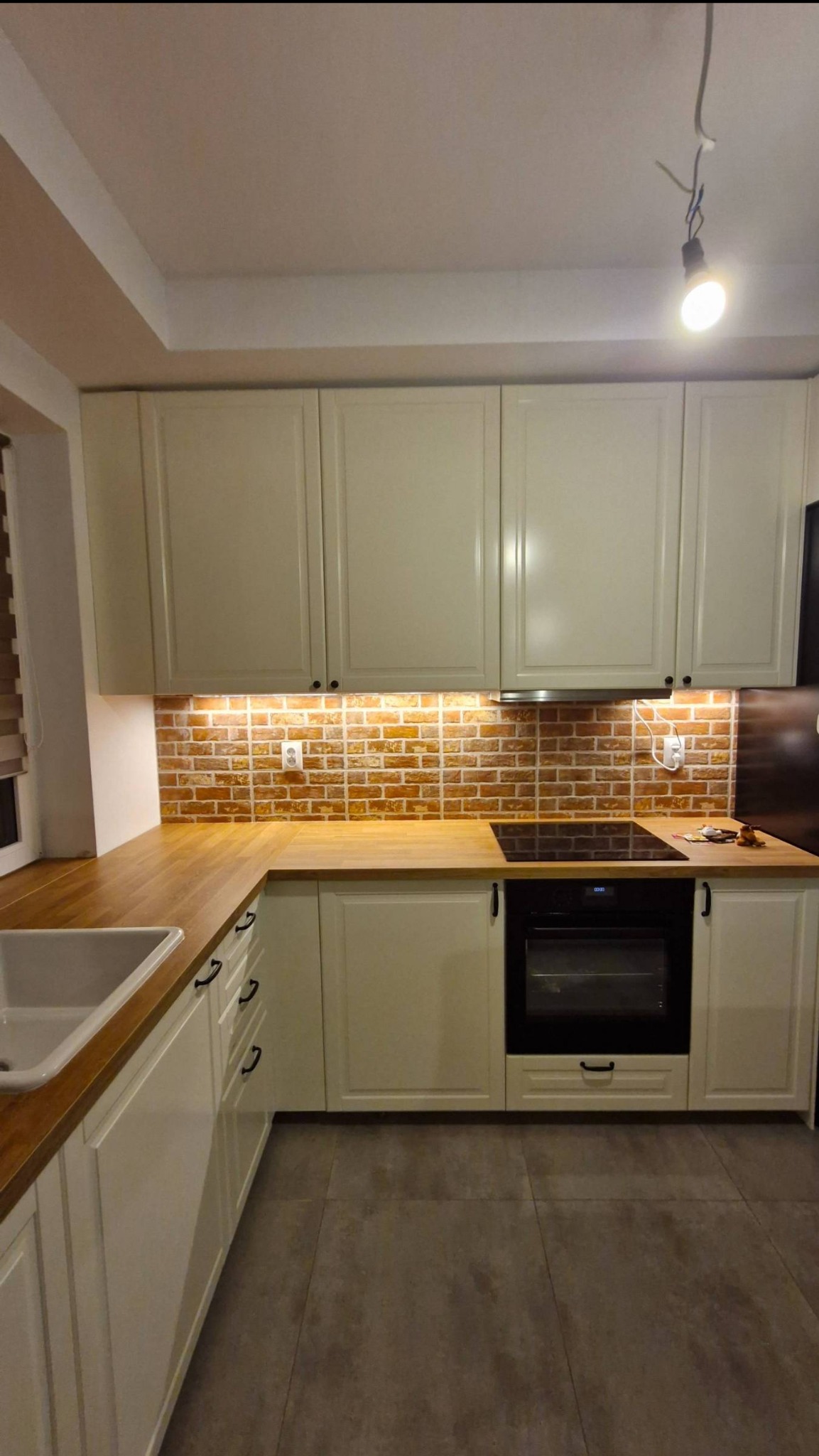 Jasna kuchnia na wymiar w Radomiu: kremowe szafki z drewnianym blatem, ceglana ściana nad blatem, czarna płyta indukcyjna i piekarnik. Oświetlenie podszafkowe.