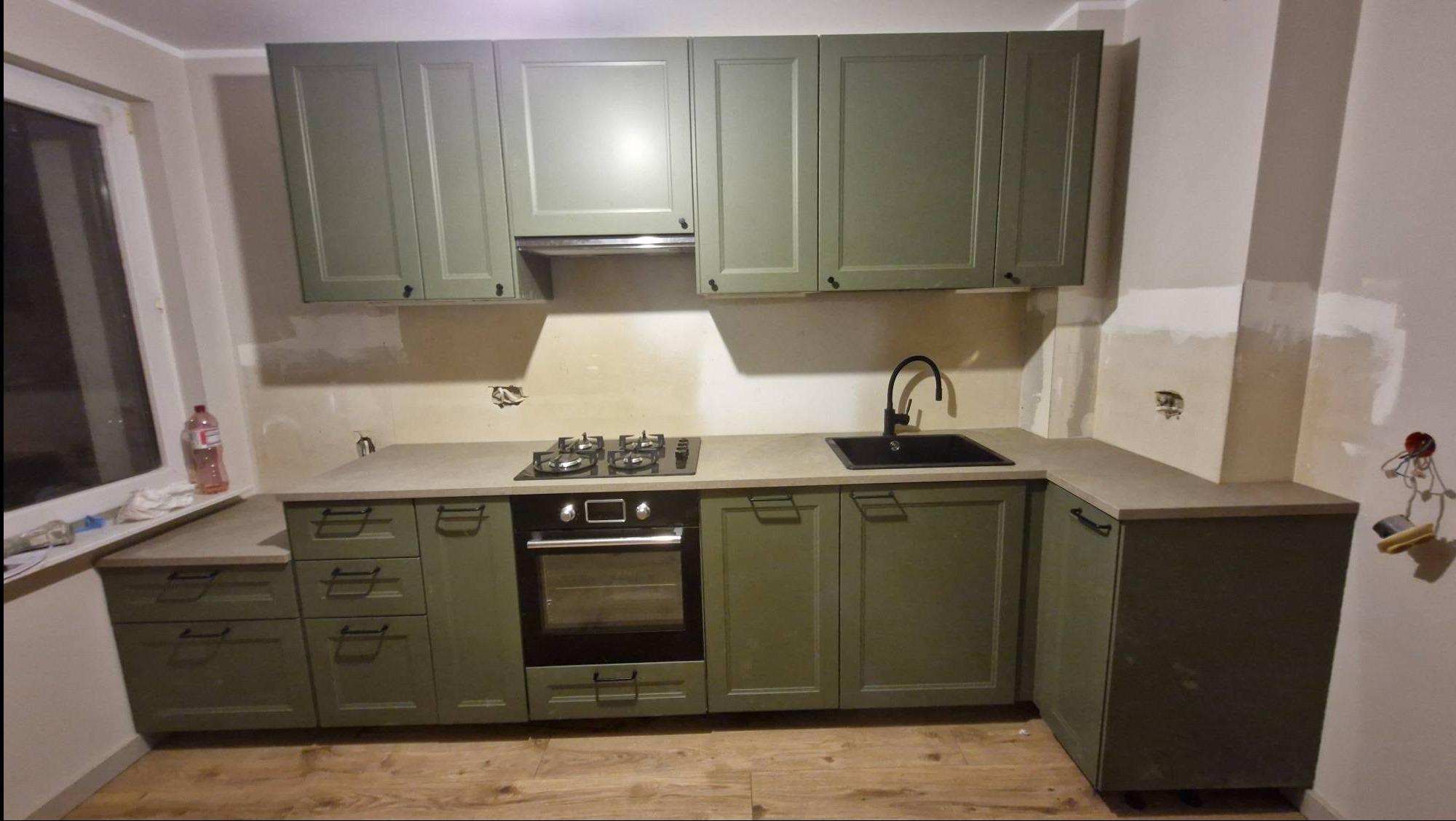Kremowo-oliwkowa zabudowa kuchenna z czarną armaturą i płytą gazową. Widoczne niedoskonałości na ścianach, podkreślające etap wykończeniowy. Ujęcie z poziomu podłogi.