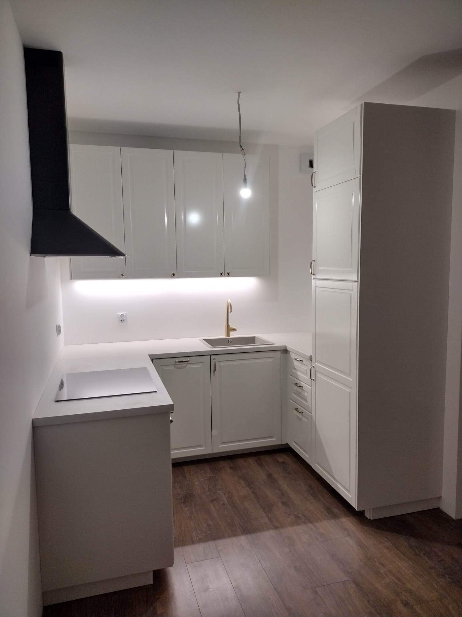 Biała, nowoczesna kuchnia z czarnym okapem i drewnianą podłogą. Uwagę przykuwa złota bateria umywalkowa i subtelne oświetlenie LED pod szafkami.