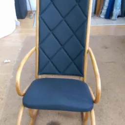 Odnowiony fotel bujany z jasnego drewna z ciemnoniebieskim, pikowanym obiciem na oparciu i siedzisku, widoczne świeże ślady po czyszczeniu na podłodze.