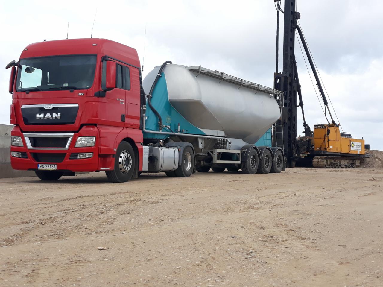 Czerwona ciężarówka MAN z naczepą-cysterną na placu budowy, w tle palownica.