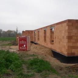 Wznoszone ściany domu z pustaków ceramicznych Porotherm, widoczne palety z materiałem budowlanym, teren budowy z błotem i zielenią, pochmurne niebo.