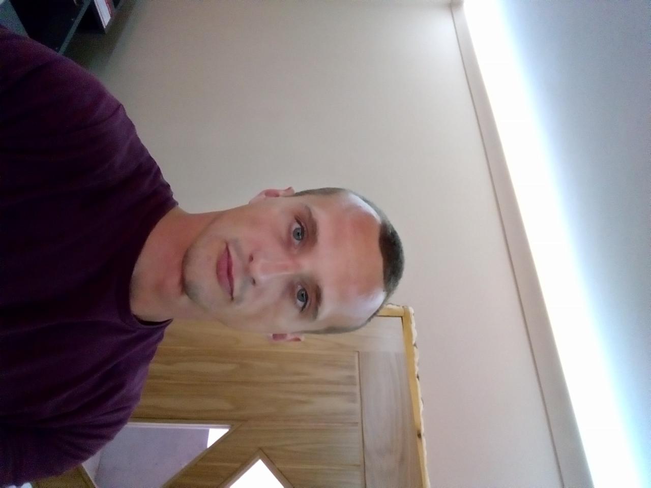 Portret mężczyzny w bordowej koszulce na tle białej ściany i drewnianych drzwi z przeszkleniem, ujęcie z dołu.