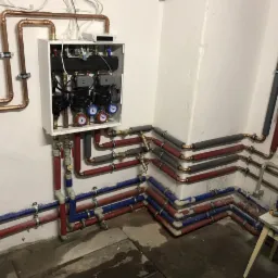 Rozbudowana instalacja grzewcza i hydrauliczna w piwnicy, z widoczną szafką rozdzielczą, rurami miedzianymi, izolowanymi i plastikowymi, z licznymi zaworami i manometrami, oraz fragmentem pieca...