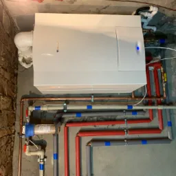 Nowoczesny piec gazowy marki Junkers zamontowany w piwnicy, widoczne rury miedziane i izolowane, filtr wody na ścianie z cegły, niebieska dioda sygnalizacyjna na piecu.