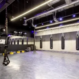 Nowoczesna sala do sportów walki z czterema workami bokserskimi wiszącymi na łańcuchach, czarną ścianą z lustrem, piłkami do ćwiczeń na półce, taśmami TRX i betonową podłogą