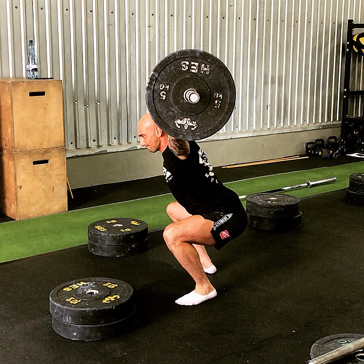 Mężczyzna z tatuażami na ramieniu wykonuje przysiad z obciążeniem w siłowni, na podłodze leżą talerze obciążeniowe, w tle skrzynia do ćwiczeń i butelka wody.