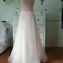 Delikatna, pudrowo-różowa suknia wieczorowa z koronkowymi aplikacjami na manekinie krawieckim, z widocznym tłem w postaci białej komody i regałów.