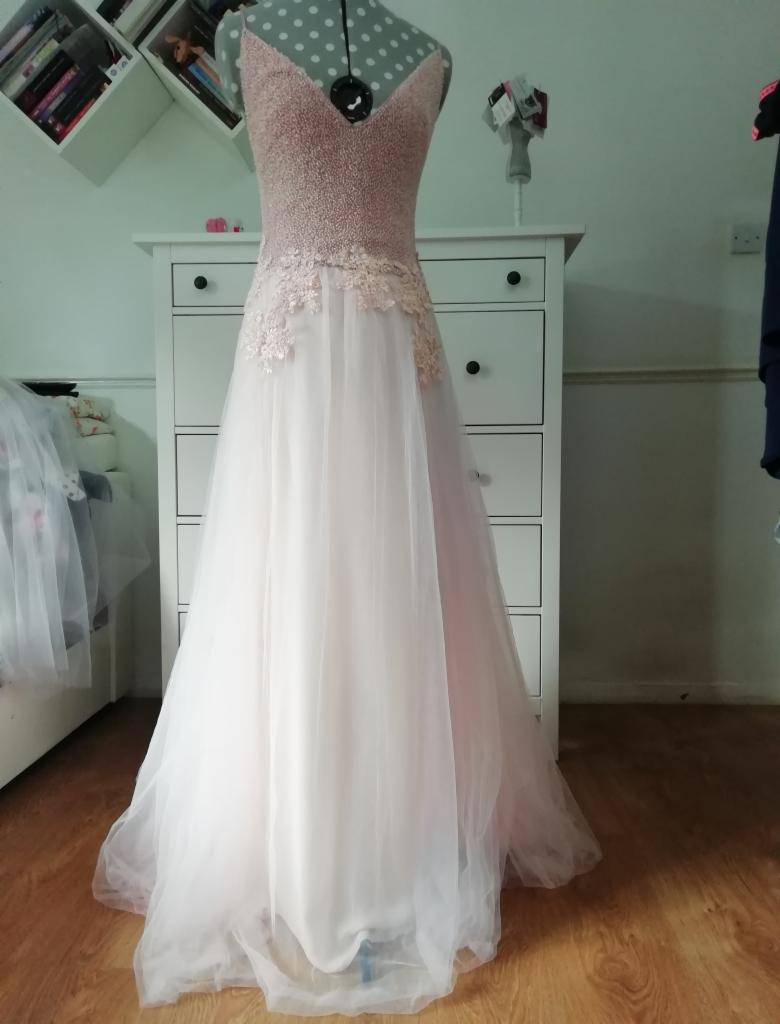 Delikatna, pudrowo-różowa suknia wieczorowa z koronkowymi aplikacjami na manekinie krawieckim, z widocznym tłem w postaci białej komody i regałów.