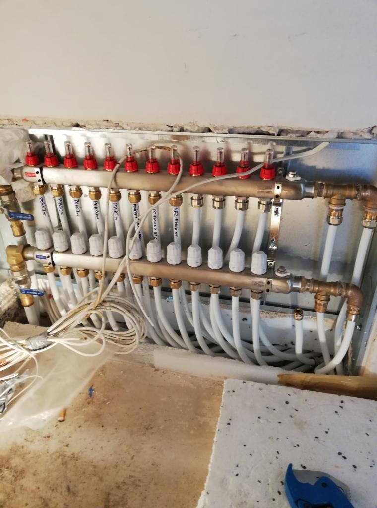 Rozdzielacz ogrzewania podłogowego z białymi rurami, zamontowany w szarej wnęce ściennej, z widocznymi zaworami i etykietami z nazwami pomieszczeń.