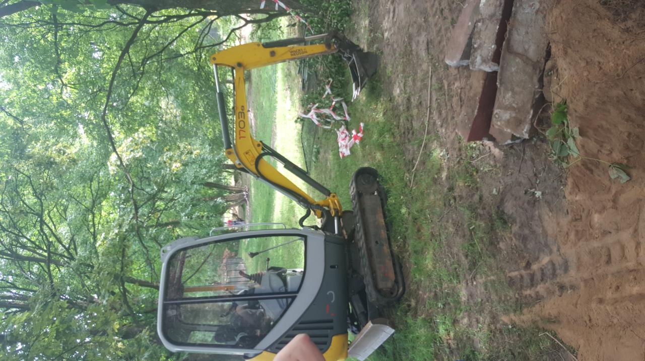 Żółto-szara mini koparka na trawiastym terenie z drzewami w tle, obok wykopu z gruzem i taśmą ostrzegawczą.