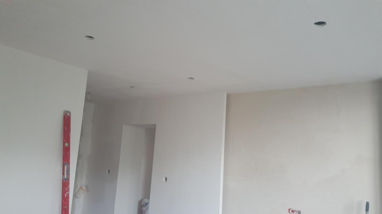 Wnętrze w trakcie prac wykończeniowych z widocznymi otworami na oświetlenie w suficie, pionową poziomicą opartą o ścianę oraz niezagruntowaną ścianą w tle.