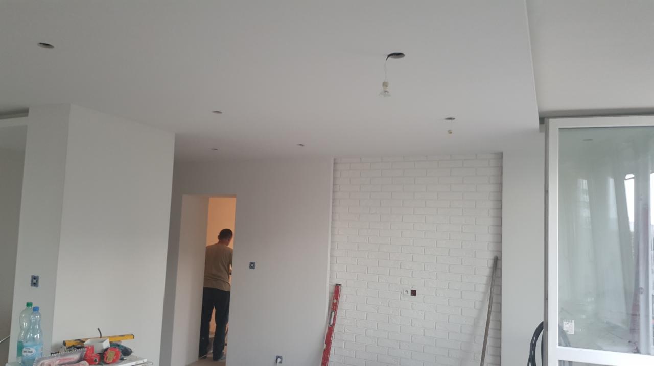 Wnętrze w trakcie remontu: białe ściany, ściana z białej cegły, widoczne narzędzia i poziomica, mężczyzna w tle w przejściu.
