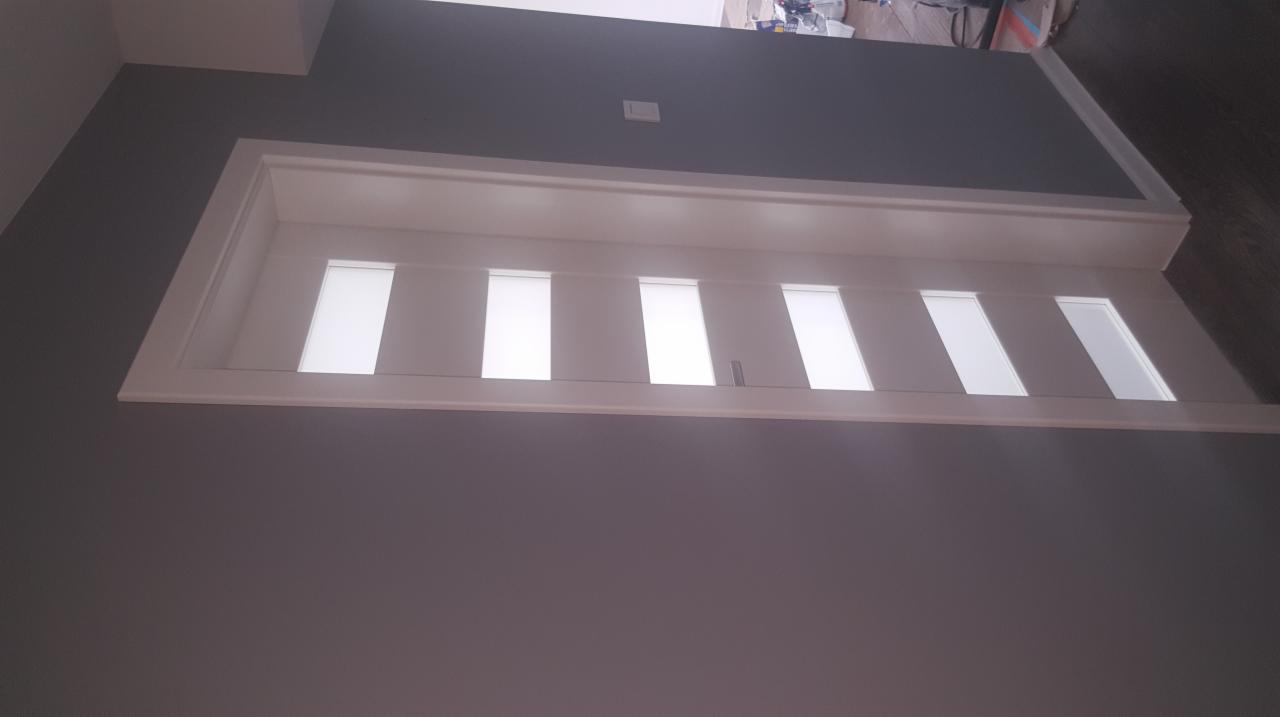 Nowoczesne, białe drzwi wewnętrzne z sześcioma przeszkleniami, wkomponowane w szarą ścianę, widoczne z dołu, w tle fragment podłogi i materiały budowlane.