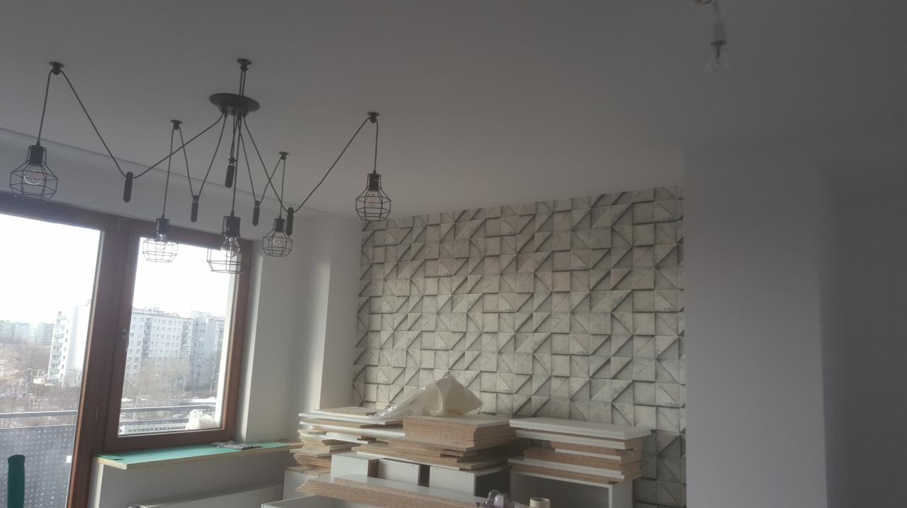 Wnętrze w trakcie prac wykończeniowych: ściana z trójwymiarową mozaiką, wisząca lampa z żarówkami w metalowych klatkach, okno z widokiem na miasto i sterta płyt meblowych w folii.