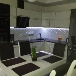 Nowoczesna, jasna kuchnia z białymi szafkami i czarnym blatem, oświetlenie LED pod szafkami, stół z ciemnymi podkładkami i roślina w doniczce, czarne krzesła z białymi oparciami.