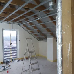 Wnętrze w trakcie montażu sufitu podwieszanego: widoczne metalowe profile, drabina, narzędzia i rura wentylacyjna owinięta folią aluminiową.