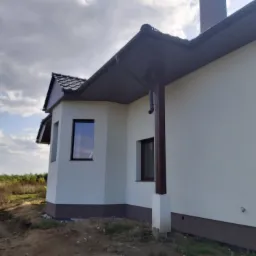 Boczny widok domu z jasną elewacją, ciemną podbitką i brązowymi ramami okiennymi, w tle pochmurne niebo i fragment krajobrazu.