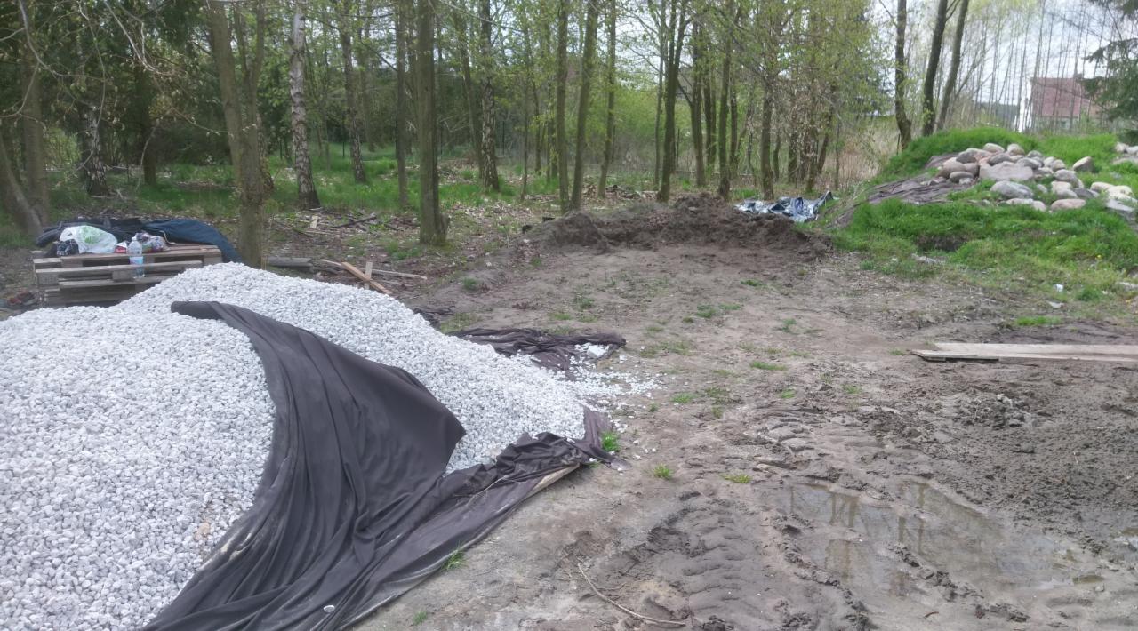 Przygotowanie terenu: stos białego kruszywa zabezpieczony czarną folią, wykopana ziemia w tle, paleta z napojami i narzędziami pod drzewami.