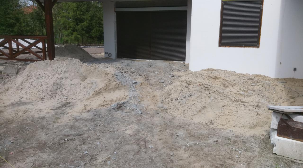 Stosy piasku i ziemi przed budynkiem z brązową bramą garażową i oknem z roletami, widoczny fragment drewnianej konstrukcji altany.