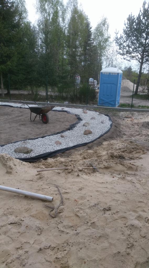 Kształtowanie ścieżki z białego żwiru i kamieni w ogrodzie, widoczna taczka i przenośna toaleta w tle.