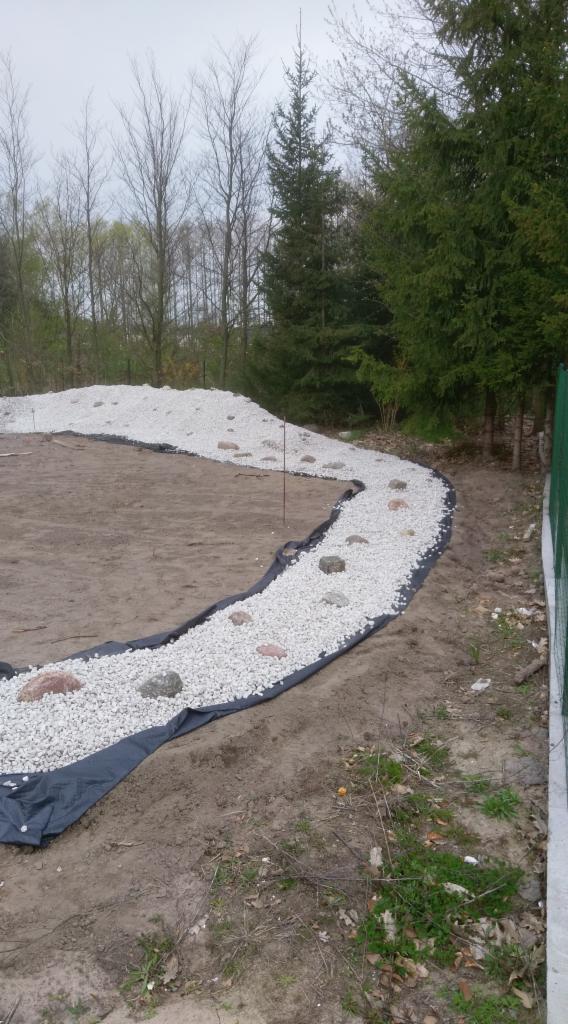 Przygotowany teren pod ścieżkę z białego grysu na czarnej geowłókninie w Bydgoszczy, widoczne większe kamienie dekoracyjne i drzewa w tle.