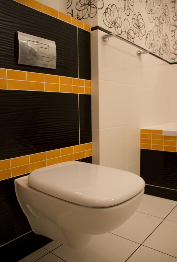 Podwieszana toaleta w łazience z czarno-białymi płytkami, akcentowanymi poziomymi pasami z żółtych płytek i wzorzystą tapetą z motywem kwiatowym.