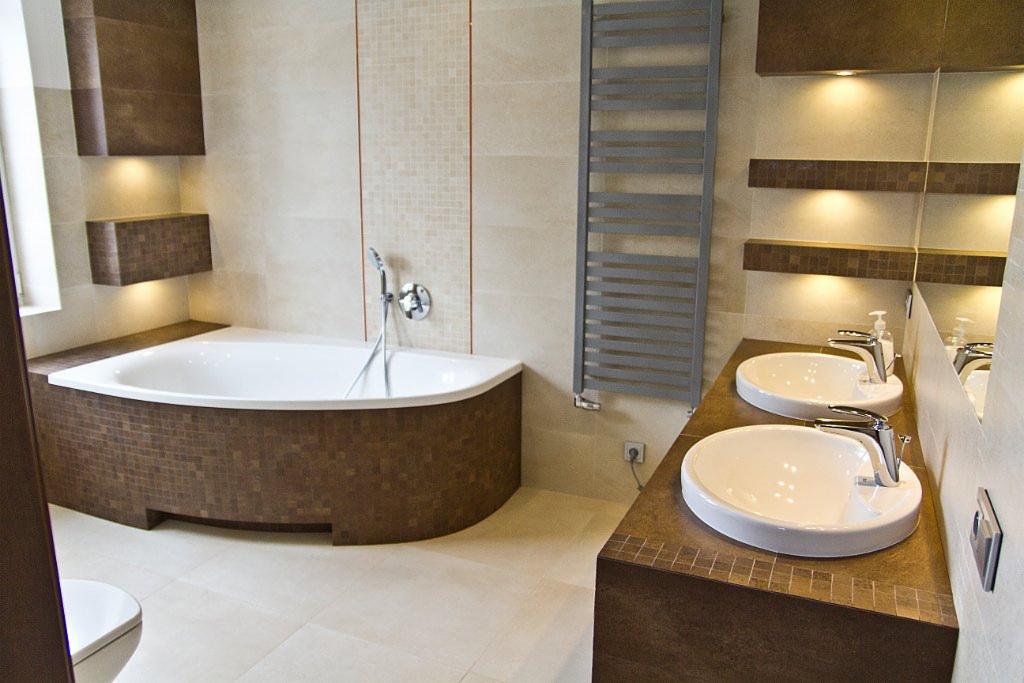 Nowoczesna łazienka z narożną wanną obudowaną mozaiką w odcieniach brązu, dwiema owalnymi umywalkami nablatowymi z chromowanymi bateriami, grzejnikiem drabinkowym i oświetleniem punktowym...