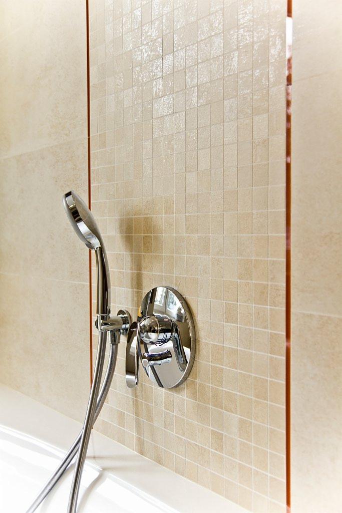 Błyszcząca, chromowana bateria prysznicowa zamontowana na ścianie wyłożonej beżowymi, kwadratowymi płytkami mozaikowymi z delikatnym połyskiem. Widoczna biała wanna i brązowa listwa dekoracyjna.