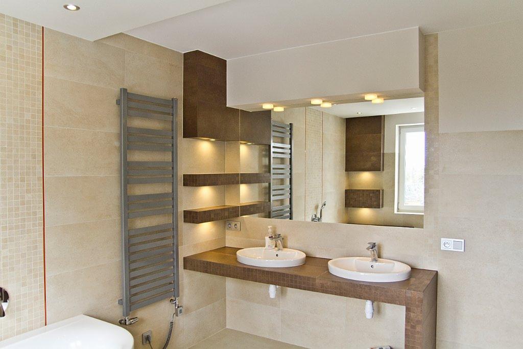 Jasna łazienka z beżowymi płytkami, dwoma umywalkami na brązowym blacie, grzejnikiem drabinkowym i oświetleniem punktowym pod półkami.