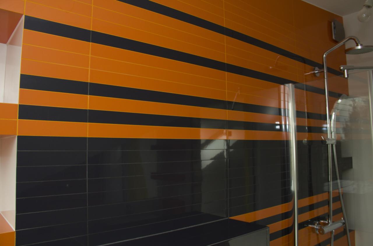 Nowoczesna łazienka z pomarańczowo-czarnymi poziomymi pasami z płytek, widoczna szklana kabina prysznicowa i chromowana bateria.