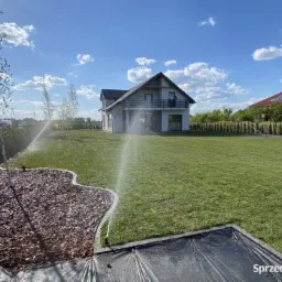 Automatyczny system nawadniania trawnika w ogrodzie z nowoczesnym domem w tle, widoczne zraszacze i świeżo nawodniona trawa.