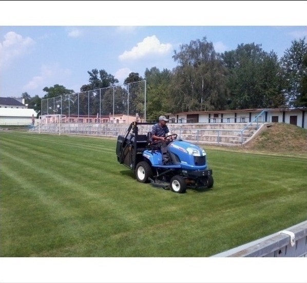 Operator na niebieskiej kosiarce samojezdnej z czarnym koszem tnącym, koszący trawę na boisku sportowym z trybunami w tle.