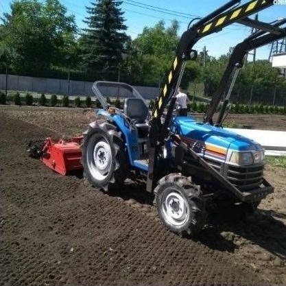 Niebieski traktor z czerwonym glebogryzem przygotowuje ziemię pod trawnik, widoczne świeżo spulchnione bruzdy w ziemi. W tle rząd małych drzewek.