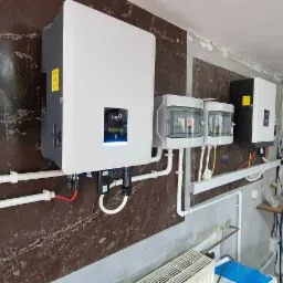 Instalacja fotowoltaiczna w pomieszczeniu: biały inwerter FOXO, rozdzielnica NOARK i zabezpieczenia na ścianie z brązową okładziną, widoczne przewody i elementy montażowe.