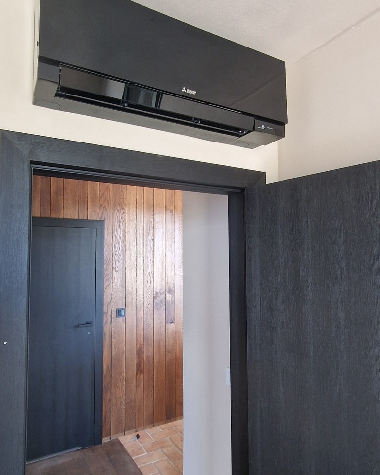 Czarny klimatyzator Mitsubishi Electric zamontowany na ścianie nad drzwiami w pomieszczeniu z boazerią i ceglaną podłogą.