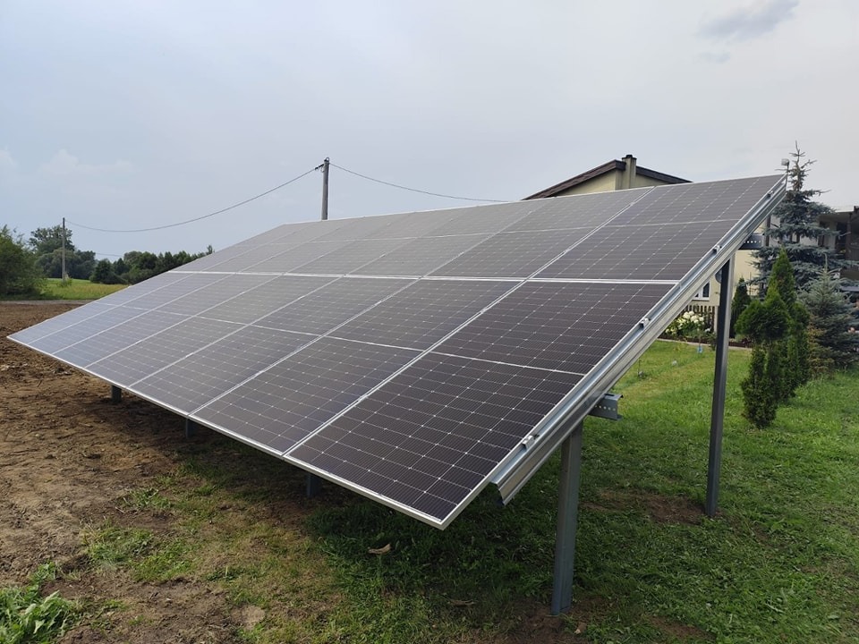 Panel fotowoltaiczny zamontowany na metalowej konstrukcji, widok z boku na trawniku przed domem w pochmurny dzień.