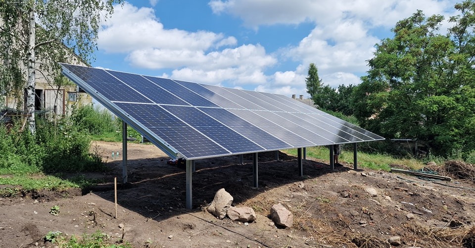 Panel fotowoltaiczny zamontowany na metalowej konstrukcji na zewnątrz, widoczne elementy montażowe, w tle budynek gospodarczy i drzewa.